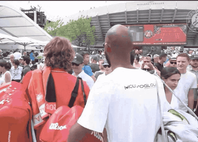 a man wearing a white shirt that says mousoglou stands in a crowd of people