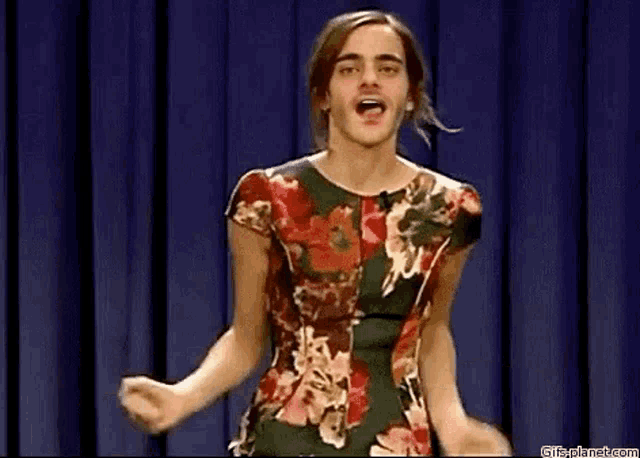 a man in a floral dress is standing on a stage with his arms outstretched .