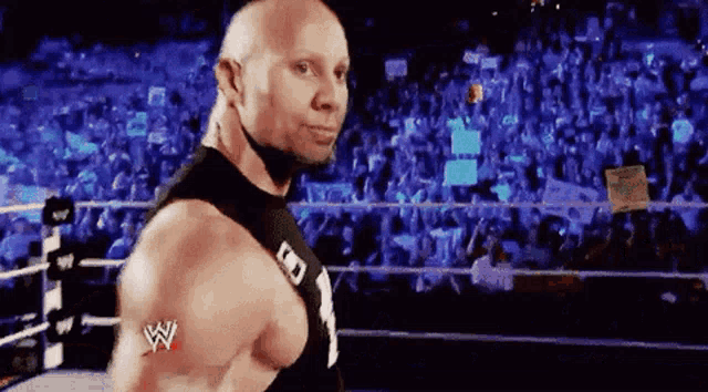 a bald wrestler is standing in a wrestling ring with a crowd behind him .