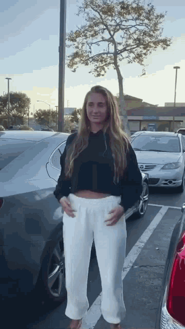 a woman in white pants and a black sweater is standing in a parking lot