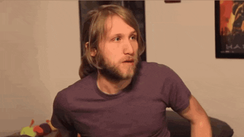 a man with a beard wearing a purple shirt with a halo poster behind him