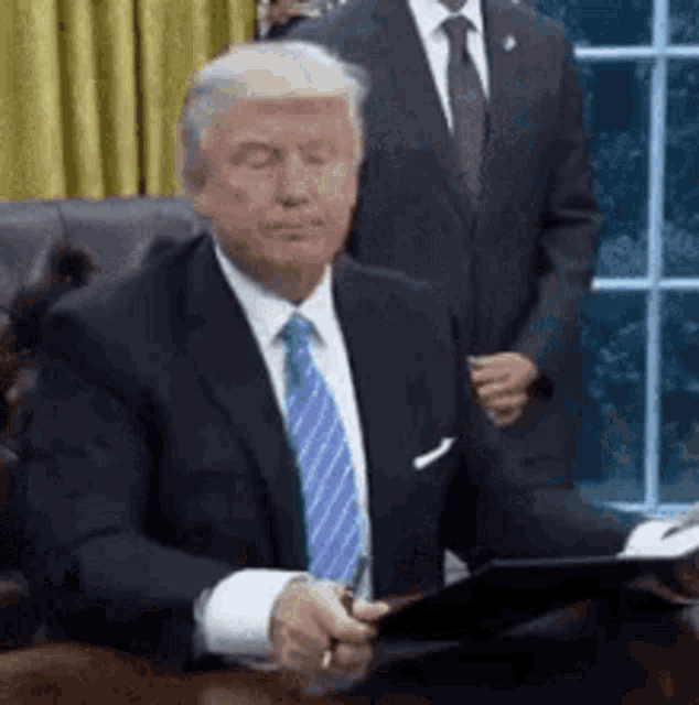 donald trump is sitting at a desk with his eyes closed while holding a tablet .