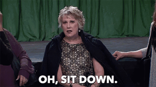 a woman in a black jacket is sitting on a couch and saying `` oh , sit down '' .