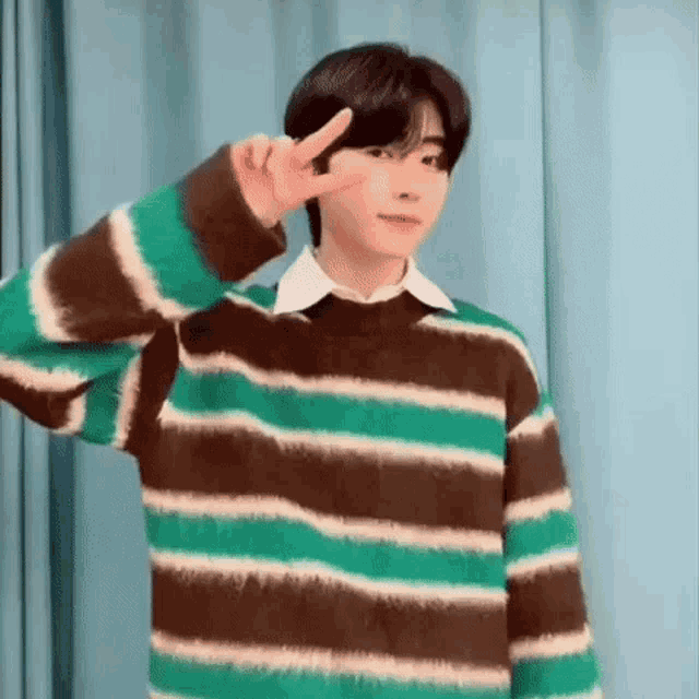 a young man is wearing a green and brown striped sweater and giving a peace sign .