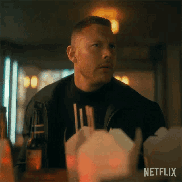 a man in a black jacket is sitting at a table with netflix written on the bottom .