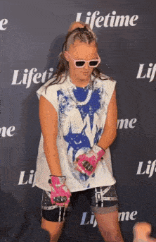 a girl wearing sunglasses and pink gloves is standing on a red carpet .