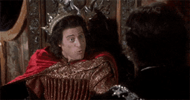 a man in a red cape is sitting on a throne and laughing while another man looks on .
