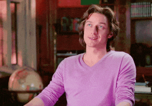 a man in a purple shirt is smiling in front of a bookshelf