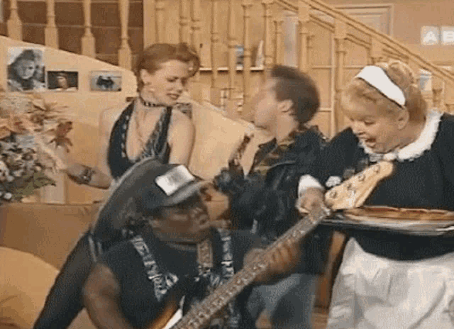 a group of people are standing around a man playing a guitar while a woman holds a tray of pizza .