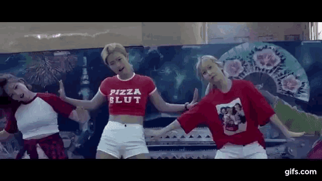 three women are dancing together and one of them is wearing a red shirt that says `` pizza slut '' .