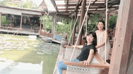 a woman sitting on a swing next to another woman in front of a pond