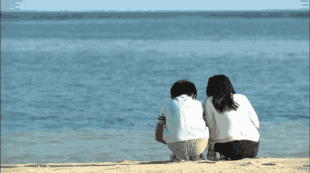 a boy and a girl sit on a sandy beach looking at the ocean