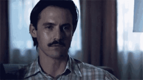 a man with a mustache is sitting on a couch in front of a window in a room .