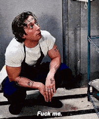 a man in an apron is squatting down in front of a metal shelf and says `` fuck me '' .