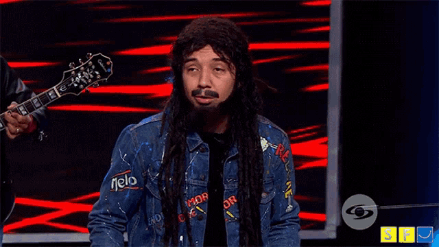 a man with dreadlocks and a beard is holding a guitar and wearing a denim jacket that says nelo on the front