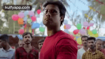 a man wearing glasses and a red shirt is standing in front of a crowd of people with balloons .