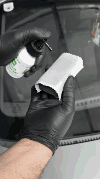 a person wearing black gloves is cleaning a windshield with a bottle of glass sealant