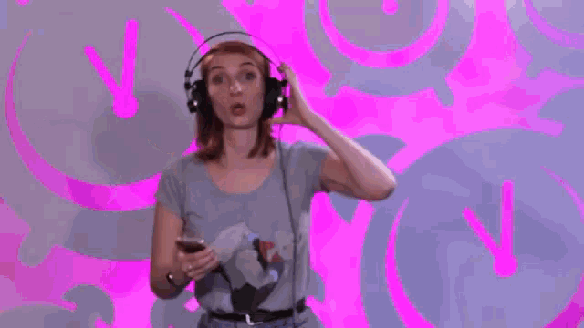 a woman wearing headphones is smiling and pointing at something in front of a pink background .