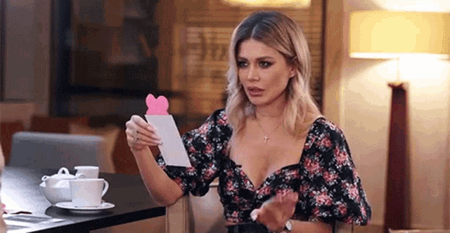 a woman in a floral dress is sitting at a table holding a card with a pink heart on it .