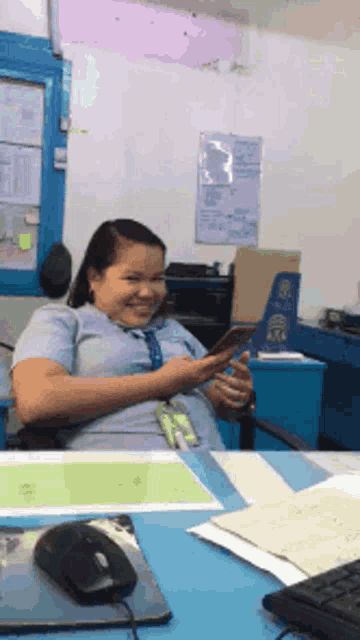 a woman is sitting at a desk looking at her phone and smiling