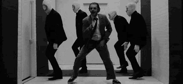 a black and white photo of a man in a suit and tie dancing with a group of men in masks .