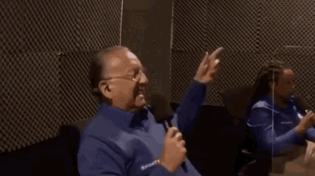 a man in a blue shirt is sitting in a chair with his hands in the air while a woman sits behind him .