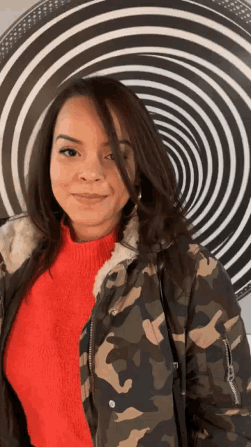a woman wearing a camouflage jacket and a red sweater is standing in front of a spiral