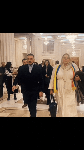 a man in a suit and a woman in a white dress are walking in a hallway