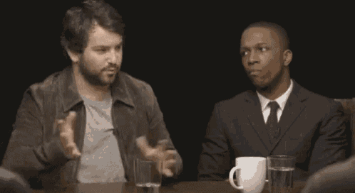 two men are sitting at a table with a cup of water and a cup of coffee .