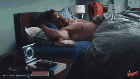 a shirtless man is laying in bed with a tecate logo on the bottom