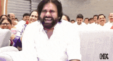 a man with a beard is laughing in front of a group of people with hk written in the corner