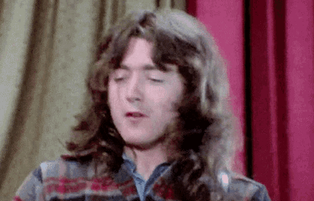 a man with long hair is standing in front of a red curtain and making a funny face .