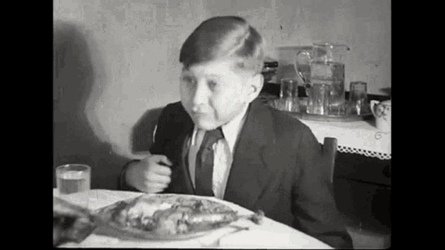 a young boy in a suit and tie is sitting at a table with a plate of food and a glass of water .