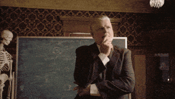 a man in a suit stands in front of a blackboard with a skeleton behind him