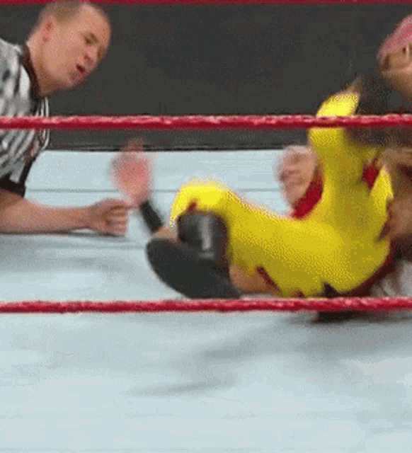 a man in a yellow suit is laying on the ground in a wrestling ring with a referee