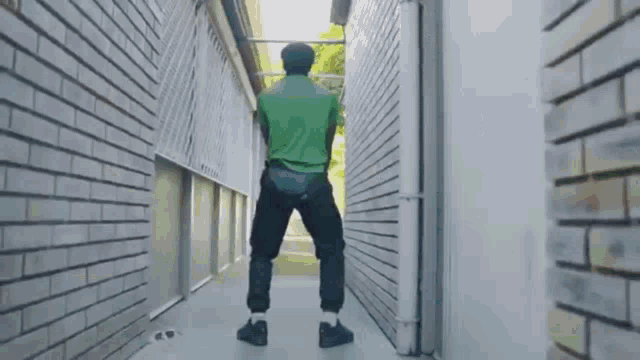 a man in a green shirt and black pants is standing in a narrow hallway between two brick buildings .