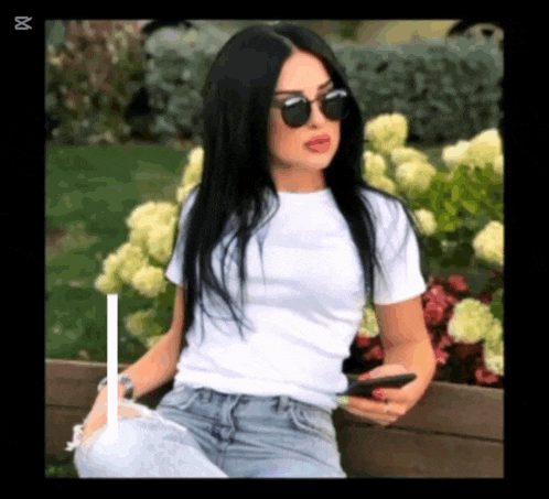 a woman wearing sunglasses and a white t-shirt is sitting on a bench .