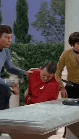 a group of people are standing around a table with a man in a red shirt sitting on top of it .