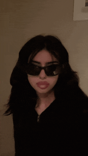 a girl wearing sunglasses and a black jacket looks at the camera with her eyes closed