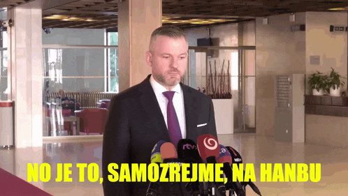 a man in a suit and tie stands in front of microphones with the words no je to samozrejme na hanbu written in yellow
