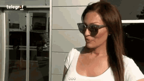a woman wearing sunglasses and a white shirt is standing in front of a glass door .