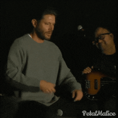 a man in a grey sweater is standing in front of a microphone in a dark room .
