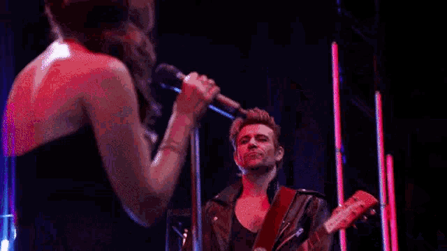 a woman is singing into a microphone on a stage while a man plays a guitar .