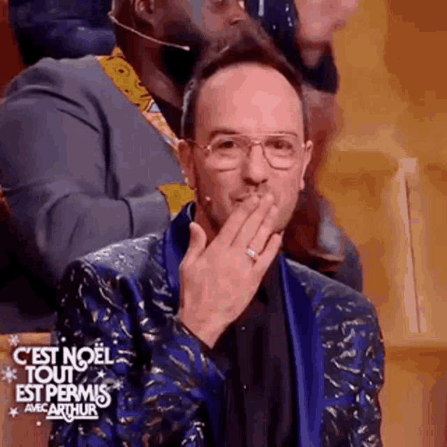 a man in a blue suit and glasses is blowing a kiss while sitting in a audience .