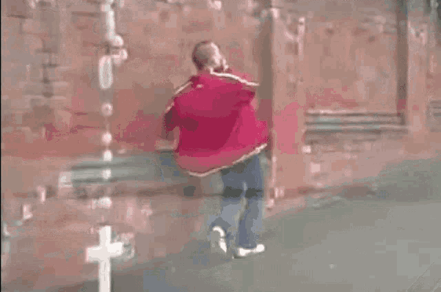 a man in a red jacket is walking down a sidewalk next to a brick wall .