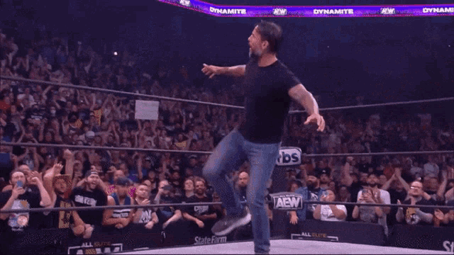 a man in a black shirt and jeans stands in a wrestling ring with a crowd watching