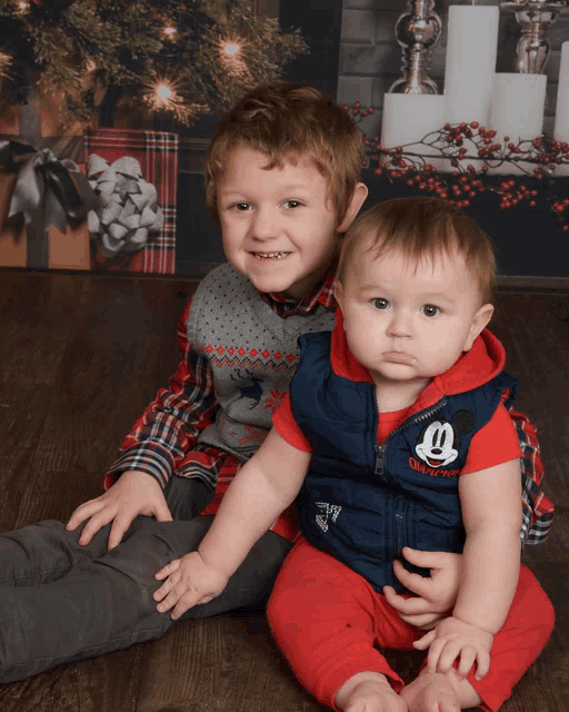 a baby wearing a mickey mouse vest sits next to a young boy wearing a plaid shirt