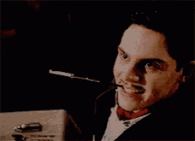 a man in a tuxedo and bow tie is smoking a cigarette and pointing at something .