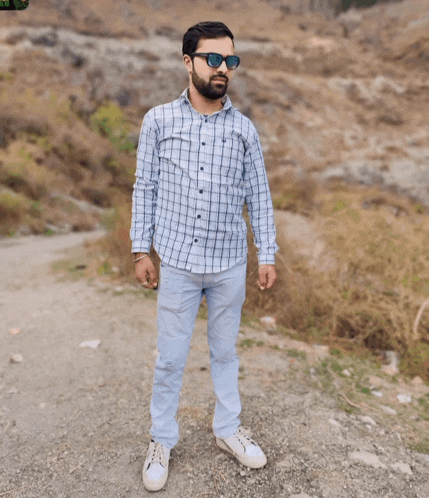 a man wearing sunglasses and a plaid shirt stands on a gravel road