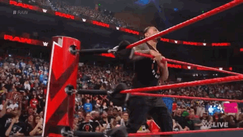 a man in a black tank top stands in a wrestling ring with a crowd behind him